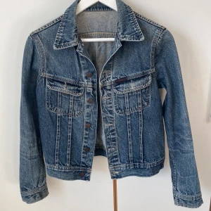 Blå jeansjacka från Rocky Denim - Fin och fräsch jeansjacka från Rocky Denim med klassisk design. Jackan har knappar framtill och två bröstfickor med lock. Lappen med storlek är borta, men ca S/M. Axelbredd 41, längd 57