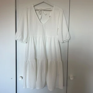 Vit klänning med knyt i ryggen - Säljer en söt vit klänning med v-ringning och knyt i ryggen. Klänningen har en luftig passform med volanger. Perfekt för varma dagar och till studenten!💕
