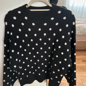 Svart stickad tröja med vita prickar - Säljer en svart stickad tröja med vita prickar. Tröjan har långa ärmar och en rund halsringning. Perfekt för kyliga dagar och ger en lekfull touch till din outfit.