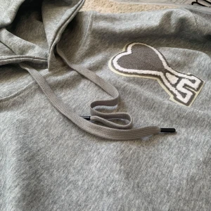 Grå hoodie från Ami Paris - Säljer en stilren grå hoodie från Ami Paris med en stor hjärtapplikation på bröstet. Tröjan har en klassisk känguruficka och justerbar huva med dragsko. Perfekt för en avslappnad look. Helt ny, aldrig använd