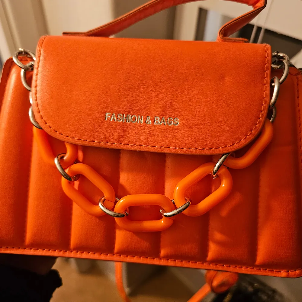 Snygg orange axelväska från Fashion & Bags med en cool kedjedetalj framtill. Väskan har en quiltad design och ett praktiskt handtag för enkel bärning. Perfekt för att ge en färgklick till din outfit! Den har lång axelrem, aldrig använt . Laukut & Käsilaukut.