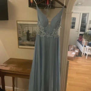 Ljusblå balklänning med spetsdetaljer - Elegant ljusblå balklänning med djup v-ringning och vackra spetsdetaljer på bysten. Klänningen har en öppen rygg med snörning och tunna axelband som knyts på ryggen.