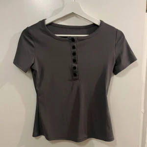 Grå kortärmad topp med knappar - Säljer en grå polyestertopp med korta ärmar och knappar framtill. Passar bra till jeans eller kjol.