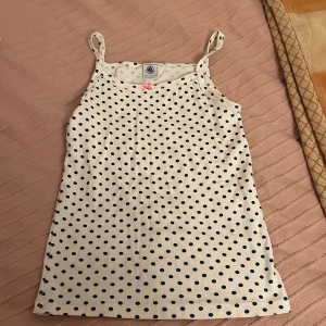 Vit prickig linnetopp från Petit Bateau - Söt vit linnetopp från Petit Bateau med mörkblå prickar och en liten rosa rosett framtill. Perfekt för varma sommardagar. Linnet har smala axelband och en bekväm passform.