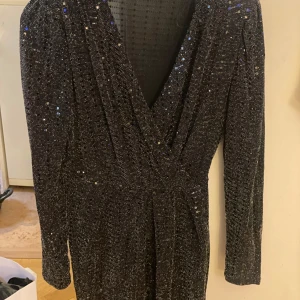 Svart glittrig klänning från Vila  - Snygg svart klänning från vila  med glittriga detaljer. Klänningen har en omlott-design och långa ärmar, perfekt för en festlig kväll. Den djupa v-ringningen ger en elegant touch.