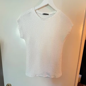 Vit topp från Zara - Säljer en stilren vit topp från Zara med en texturerad yta. Toppen har korta ärmar och en rund halsringning, perfekt för en enkel och elegant look. Använd gärna köp nu🌸