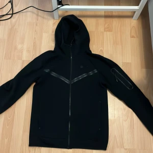 Svart hoodie från Nike - Säljer en stilren svart hoodie från Nike med dragkedja och huva. Jackan har långa ärmar och en diskret logga på bröstet. Perfekt för en sportig look eller avslappnade dagar.