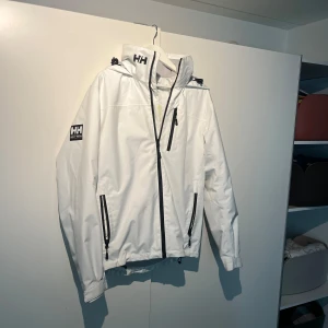 Vit vindjacka från Helly Hansen - Säljer en stilren vit vindjacka från Helly Hansen med justerbar huva och dragkedja framtill. Jackan har svarta detaljer och logotyp på ärmen och axeln. Perfekt för blåsiga och regniga dagar.