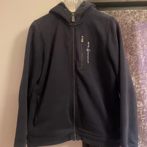 Mörkblå jacka från Sail Racing - Säljer en mörkblå jacka från Sail Racing med dragkedja och huva. Jackan har en stilren design med logotyp på bröstet och en praktisk bröstficka med dragkedja. Perfekt för kyligare dagar. Ingen Skador, Bra skick.