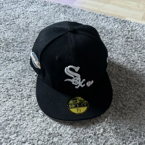 Svart keps från New Era - Snygg svart keps från New Era med broderad logga framtill. Kepsen har en platt skärm och är perfekt för en sportig look. Den har även en patch på sidan som ger extra stilpoäng.