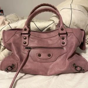 Rosa axelväska med dragkedja - Snygg rosa axelväska i skinn med flätat handtag och flera dekorativa nitar. Den kan användas som skolväska eller vanlig handväska. Fin och i bra skick. Pris kan alltid diskuteras vid snabb affär! Mått: 40cm i längd, 25cm i höjd och 10 cm i bredd.  Axelrem medföljder! 