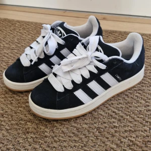 Svarta Adidas Campus sneakers säljs billigt! Köpt för 1 år sedan men använts bara fåtal gånger.  - Snygga svarta Adidas Campus sneakers med vita ränder och breda snören. Köpta för 1 år sedan men har bara använts några få gånger. Skorna har en klassisk design med en bekväm passform och en gummisula som ger bra grepp. Perfekta för en avslappnad stil. 