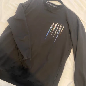 Svart tröja från Amiri med färgglatt tryck - Säljer en svart tröja från Amiri med ett coolt färgglatt tryck som rinner ner från loggan. Tröjan har långa ärmar och ribbade muddar vid ärmslut och nederkant. Skriv för fler frågor och bilder🎀
