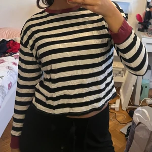 Randig långärmad tröja med vinröda detaljer - Säljer en svartvit randig långärmad tröja med vinröda muddar och halsringning. Perfekt för en avslappnad stil. Passar bra till jeans eller kjol.