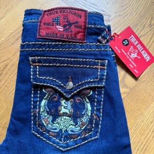Blå jeans från True Religion - Snygga blå jeans från True Religion med kontrastsömmar och broderad bakficka. Jeansen har en klassisk femficksdesign och en knappgylf. Perfekta för en stilren look. Observera att jeansen är low waist