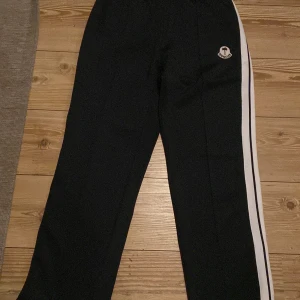 Svarta byxor med vita ränder från Moncler - Snygga svarta byxor från Moncler med vita ränder längs sidorna. Byxorna har en broderad logga på framsidan och dragkedjor vid bensluten. Perfekta för en stilren look. Storleken är XL men passar som L