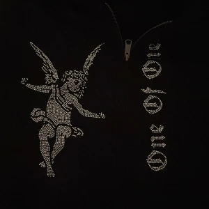 Svart hoodie från One of One (XL) - Säljer en svart hoodie från One of One med ett  ängelmotiv i stenar på framsidan. Hoodien har en dragkedja och texten 'One of One' i samma stil. Hoodie har aldrig använts och är som ny. Den är i storleken XL och har en bra passform. 