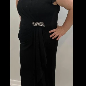 Svart glittrig klänning med dekorativ detalj - Elegant svart klänning med glittrande detaljer och en dekorativ spänne i midjan. Klänningen har en omlott-design och korta ärmar, perfekt för en festlig kväll.