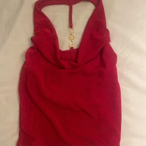 Röd halterneck topp med gulddetaljer - Snygg röd halterneck topp med dekorativa gulddetaljer och strass på ryggen. Perfekt för en kväll ute eller en fest. Toppen har en draperad framsida och är tillverkad i ett mjukt material.