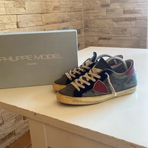 Sneakers från Philippe Model - Snygga sneakers från Philippe Model i en mix av grått och rött med vita snören. Skorna har en klassisk design med en bekväm sula och är perfekta för en stilren look.