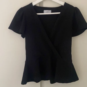 Svart omlottopp från Holly & Whyte - Snygg svart omlottopp från Holly & Whyte med korta ärmar och en elegant v-ringning. Perfekt för en stilren look. Toppen har en lätt volangdetalj vid nederkanten som ger en feminin touch. Jätte fin nu till sommaren, Använt den 1-2 gånger.