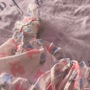 Blommig offshoulder topp - Säljer en söt blommig offshoulder topp med volangdetaljer och knyt framtill. Toppen har ett romantiskt mönster i rosa, blått och vitt. Perfekt för varma sommardagar! Köpt i Thailand och är helt ny