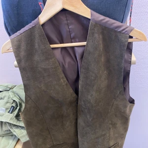 Brun mockaväst - Snygg brun mockaväst med v-ringad design. Perfekt för en stilren look. Västen har en slät yta och är fodrad på insidan för extra komfort.