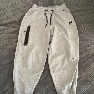 Grå mjukisbyxor från Nike - Snygga grå mjukisbyxor från Nike med svart snörning och praktisk ficka på sidan. Perfekta för en avslappnad stil. Byxorna har elastisk midja och Nike-logga på benet.