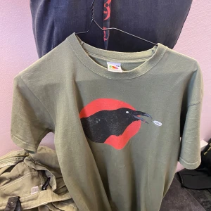 Olivgrön t-shirt med queens of stone age tryck från Fruit of the Loom - Säljer en olivgrön t-shirt med ett coolt tryck av en korp och röd cirkel. På baksidan finns texten 'Queens of the Stone Age'. Perfekt för musikälskare! 🎸