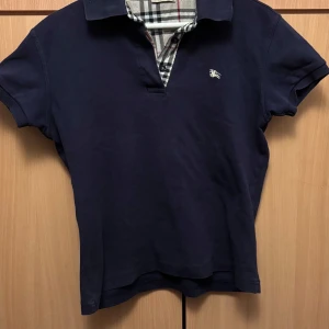 Mörkblå kortärmad t-shirt från Burberry - Säljer en snygg mörkblå kortärmad t-shirt från Burberry. Det står att den är i XL men passar storlek S.. Den har en stilrent rutigt mönster på insidan och en liten broderad logga på bröstet. Tillverkad i 100% bomull för en bekväm känsla. Perfekt för en stilren look! 