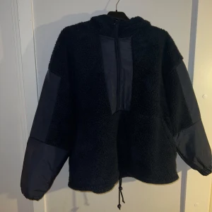 Mörkblå sportjacka/anorak - Mysig mörkblå teddyjacka med dragkedja framtill och huva. Jackan har långa ärmar med glansiga detaljer och en loose passform. Perfekt för kyliga dagar. Man kan justera hur tight man vill att jackan ska sitta vid höfterna. Använd 1-2 gånger så i nyskick. Oversized