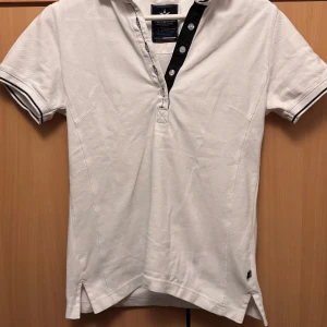 Vit kortärmad pikétröja från Texstar - Säljer en stilren vit pikétröja från Texstar i storlek S. Tröjan har korta ärmar med svarta detaljer och knappar framtill. Tillverkad i 100% bomull för en bekväm känsla. Perfekt för en avslappnad look.