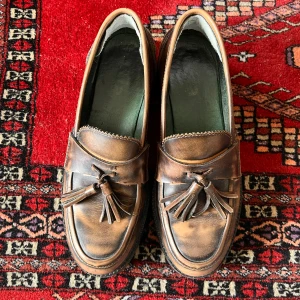 Bruna loafers i läder med tofsar strl 38 - Bruna loafers i läder med tofsar i strl 38. Kommer med hälgrepp insatta, går att ta av. Snygg fade på skorna och är i bra skick. Ingen kartong!