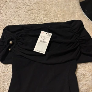 Svart offshoulder topp med pärldetalj - Trendig svart offshoulder topp med ribbad kropp och snygga rynkade ärmar. Toppen har dekorativa pärlor på sidorna och sitter tight över axlarna. Perfekt för dig som vill ha en stilren men ändå unik look.