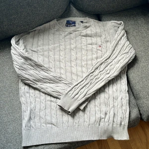 Ljusgrå kabelstickad tröja från Gant storlek M - Kabelstickad tröja från Gant i ljusgrått med rund hals och diskret broderad logga på bröstet. Tröjan har ribbade muddar och nederkant, samt långärmad passform. Perfekt för dig som gillar klassisk och stilren look.
