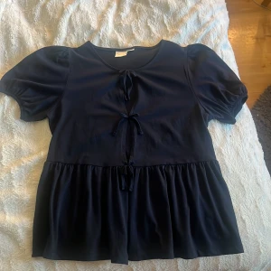 Mörkblå blus med knyt från Gina Tricot - Supersöt mörkblå blus från Gina Tricot med puffiga korta ärmar och tre knyt framtill. Blusen har en lätt utställd volang nedtill och rund halsringning. Har aldrig använt men prislappen tog jag bort när jag köpte den! 
