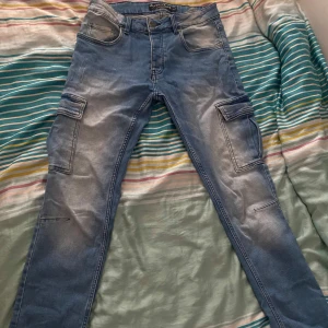 Blå cargopants från Amaci.de W30 L32 - Säljer ett par ljusblå cargopants från Amaci.de med coola fickor på sidorna och slitna detaljer. Byxorna har normal passform och raka ben, tillverkade i jeansmaterial. Perfekt för dig som gillar streetstyle och vill ha extra plats för prylar i fickorna.