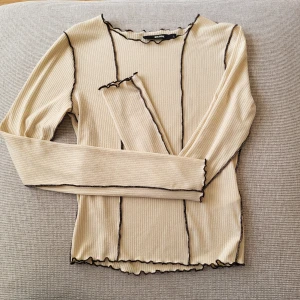 Beige ribbad långärmad topp Bik Bok - Säljer en beige ribbad långärmad topp från Bik Bok med kontrastfärgade svarta sömmar och vågiga kanter vid hals, ärmslut och nederkant. Slim fit och stretchigt material som sitter snyggt på kroppen. Mycket lite nopprig, inget jag anser förstör helhetsintrycket.