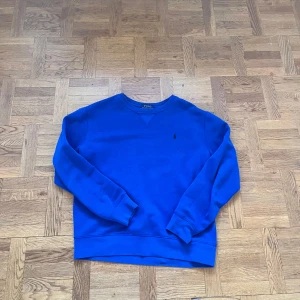 Blå sweatshirt från Polo Ralph Lauren - Snygg blå sweatshirt från Polo Ralph Lauren med klassisk rund hals och broderad logga på bröstet. Tröjan har långa ärmar och ribbade muddar vid ärmslut och nederkant. Perfekt för dig som gillar stilren och enkel design.