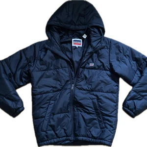 Svart pufferjacka från Levi's - Snygg Svart pufferjacka från Levi's med huva och dragkedja framtill. Jackan har quiltad design, två sidofickor och en liten logotyp på bröstet. Perfekt nu inför hösten. DEN SER MÖRKBLÅ UT MEN ÄR SVART. 