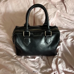 Svart handväska i skinn  - Snygg svart handväska i skinn från rizzo Väskan har två dragkedjor upptill och vita kontrastsömmar som ger en cool look. Perfekt storlek för att få plats med allt du behöver till vardags.