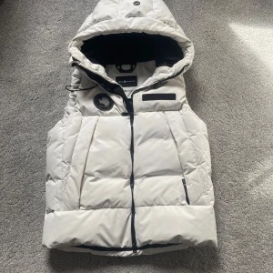 Vit dunväst med huva från sail racing  - Snygg vit dunväst från sail racing med svart dragkedja och huva. Västen har två fickor framtill och coola logodetaljer på bröstet. Perfekt för lager-på-lager och håller dig varm under kyliga dagar.