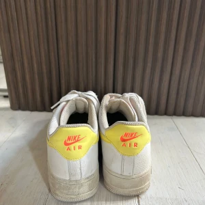 Nike Air Force 1 vita med gula detaljer - Nike Air Force 1 sneakers i vitt skinn med gula detaljer på häl och swoosh. Klassisk låg modell med snörning och perforerad tå för extra ventilation. Snygg kontrast mellan det vita och det gula, samt orange Nike-logga på hälen.