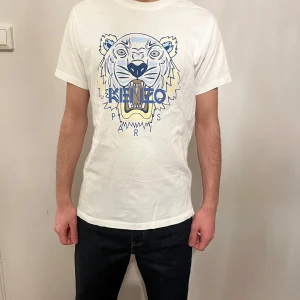 Vit Kenzo t-shirt med tigertryck - Säljer en vit t-shirt från Kenzo med ikoniskt tigertryck i blått och gult på bröstet. T-shirten har rund hals och korta ärmar, perfekt för dig som vill sticka ut med en cool design och snygg logga framtill.