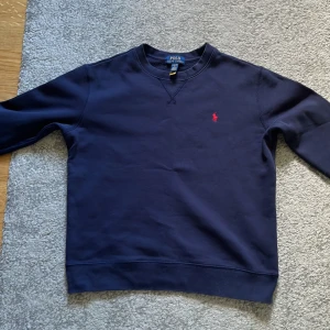 Mörkblå sweatshirt från Polo Ralph Lauren passar L barn och M ungdom  - Säljer en stilren mörkblå sweatshirt från Polo Ralph Lauren med klassisk rund hals och röd broderad logga på bröstet. Tröjan är i mjuk bomull och har långa ärmar, perfekt för en chill och snygg look.
