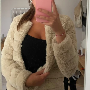 Beige teddyjacka med fluffig look - Supermjuk och fluffig beige teddyjacka med rund halsringning och diskreta knappar framtill. Jackan har en boxig passform och är perfekt för dig som vill ha en mysig och trendig stil. Passar till allt och ger en chill vibe.