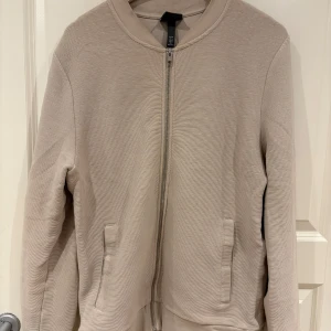 Beige bomberjacka från H&M - Snygg beige bomberjacka från H&M med diskret struktur i tyget. Jackan har dragkedja framtill, två fickor och en klassisk bomberkrage. Perfekt för dig som gillar en clean och stilren look.