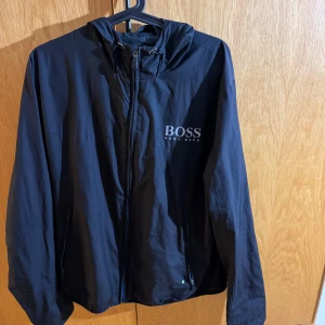 Svart vindjacka från Hugo Boss - Snygg svart vindjacka från Hugo Boss med huva och dragkedja framtill. Jackan har logga på bröstet och på huvan, samt två fickor med dragkedja. Perfekt för dig som vill ha en stilren och sportig look. Märkena är reflekterade vid ljus