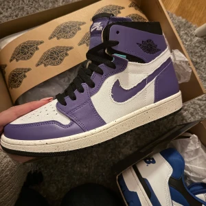 Nike Air Jordan 1 High Lila/Vit - Nike Air Jordan 1 High sneakers i lila och vitt med svart snörning och detaljer. Ovandel i skinn och textil, klassisk Jordan-logga på sidan och tungspets. Sula i vitt med svarta och lila stänk, samt lila yttersula. 