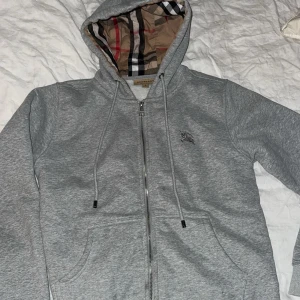 Grå hoodie från Burberry med dragkedja - Snygg grå hoodie från Burberry med klassiskt rutigt foder i huvan och broderad logga på bröstet. Tröjan har hel dragkedja, snörning i huvan och två fickor framtill. Perfekt för dig som gillar stilrena och exklusiva plagg.